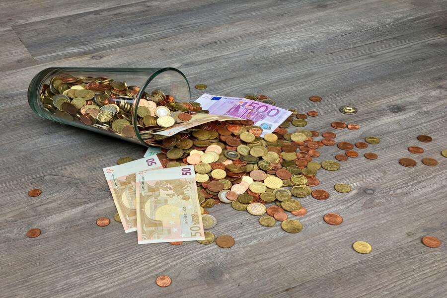 Un vaso de cristal volcado sobre un suelo de madera con monedas de euro y varios billetes de euro, entre ellos de 50 y 500 denominaciones, esparcidos por los alrededores, tal vez después de declarar cobro herencia.
