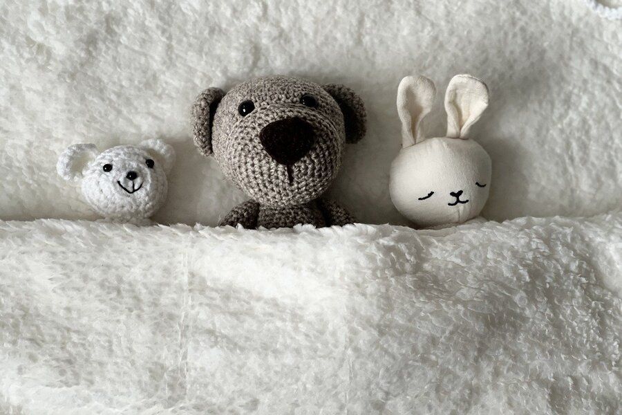 Tres peluches -un pequeño oso blanco, un oso marrón y un conejo beige- yacen uno junto al otro bajo una mullida manta blanca, acogidos como si compartieran herencia y legado sobre una superficie suave y texturizada.