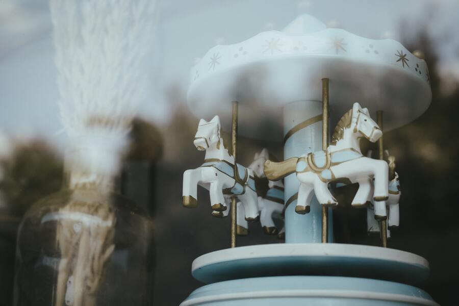 Primer plano de un pequeño carrusel decorativo con caballos blancos y palos dorados, junto a una botella que contiene trigo seco, visto a través de una ventana con reflejos, que capta el delicado encanto de la herencia yacente. Primer plano de un pequeño carrusel decorativo con caballos blancos y palos dorados, junto a una botella que contiene trigo seco, visto a través de una ventana con reflejos, que capta el delicado encanto de la herencia yacente.