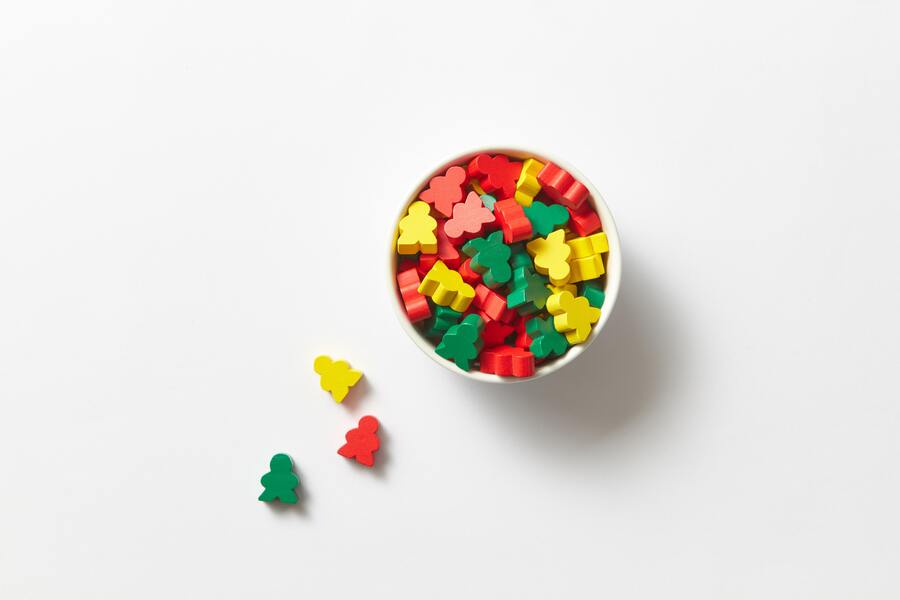 Un cuenco blanco lleno de pequeñas piezas de juego de mesa de colores con forma de personas en rojo, amarillo y verde representa herencia yacente. Tres piezas de juego se colocan fuera del cuenco sobre un fondo blanco.