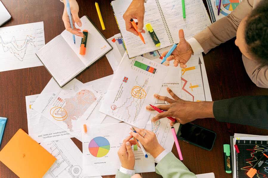 Cuatro personas se sientan alrededor de una mesa cubierta de tablas, gráficos, cuadernos y rotuladores fluorescentes, hablando de tipos de despedido y señalando documentos y notas, con un smartphone entre el material.