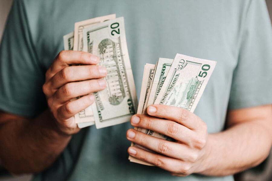 Una persona con una camisa verde claro sostiene y cuenta varios billetes de dólar estadounidense, entre ellos de 20 y 50 dólares, quizá pensando en sus finanzas tras informarse sobre los tipos de despedido.