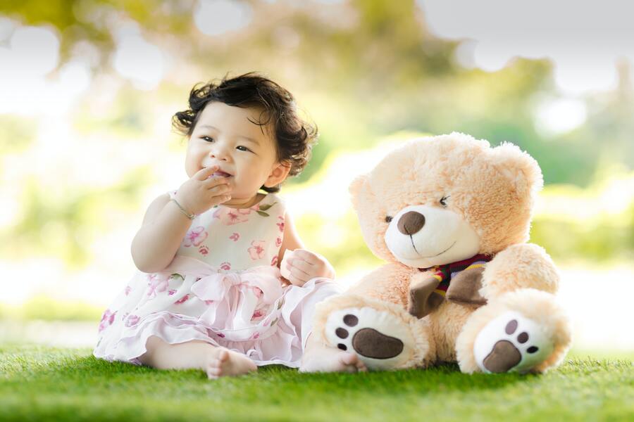 Un bebé con un vestido rosa y blanco está sentado en la hierba junto a un gran osito de peluche marrón claro, sonriendo con una mano cerca de la boca. Este dulce momento capta la alegría y la calidez de la filiación temprana, sobre un fondo de vegetación difuminada.