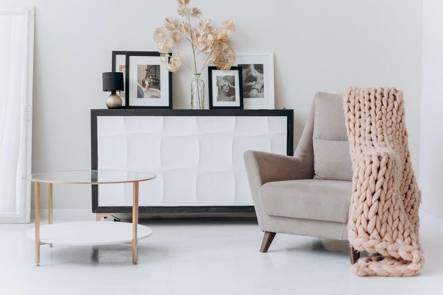Un moderno salón con un sillón beige cubierto con una gruesa manta de punto rosa, una mesa de centro redonda de cristal y un aparador con fotos enmarcadas, como un testamento del uno para el otro, además de una lámpara y plantas secas decorativas. Un moderno salón con un sillón beige cubierto con una gruesa manta de punto rosa, una mesa de centro redonda de cristal y un aparador con fotos enmarcadas, como un testamento del uno para el otro, además de una lámpara y plantas secas decorativas.