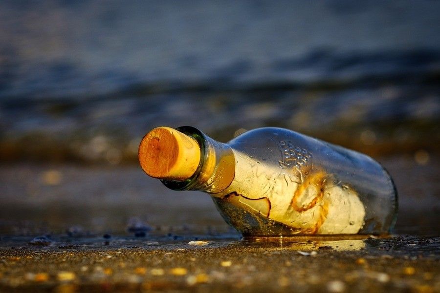 Testamento del uno para el otro: ¿Qué es y cómo funciona? Una botella de cristal cerrada con un corcho yace sobre la arena húmeda cerca de la orilla, conteniendo un trozo de papel enrollado: un posible testamento del uno para el otro. El mar y las suaves olas se ven al fondo.