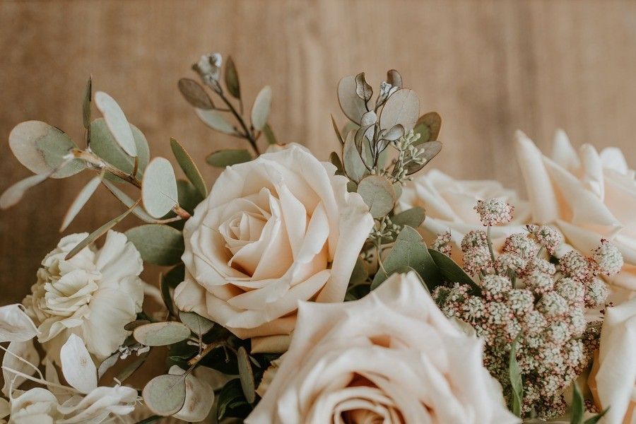Primer plano de un arreglo floral con rosas de color crema pálido, hojas de eucalipto, flores blancas y racimos de pequeños capullos rosáceos, sobre un fondo marrón neutro: cada flor es tan única como los bienes privativos.