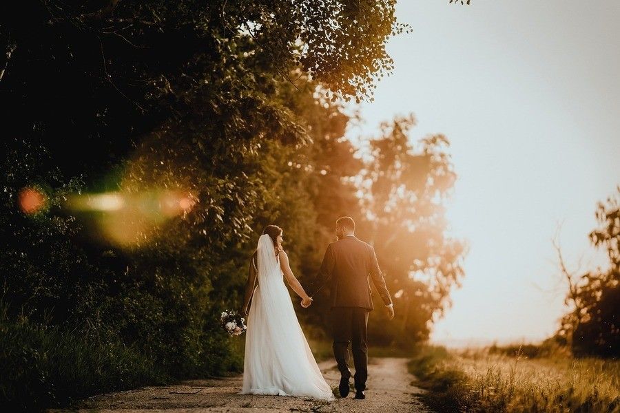 Una novia vestida de blanco y un novio, listos para casarse por lo civil en España, caminan de la mano por un sendero rural al atardecer, rodeados de árboles y bañados por la cálida y resplandeciente luz del sol. Una novia vestida de blanco y un novio, listos para casarse por lo civil en España, caminan de la mano por un sendero rural al atardecer, rodeados de árboles y bañados por la cálida y resplandeciente luz del sol.