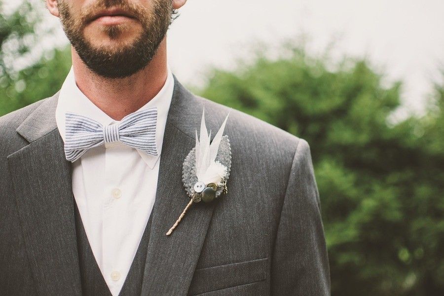 ¿Cuáles son los requisitos para casarse por lo civil en España? Un hombre con traje gris y pajarita, preparándose para casarse por lo civil en España, aparece de hombros para arriba. Lleva barba y un boutonniere con plumas blancas y detalles verdes en la solapa, sobre un fondo verde difuminado.