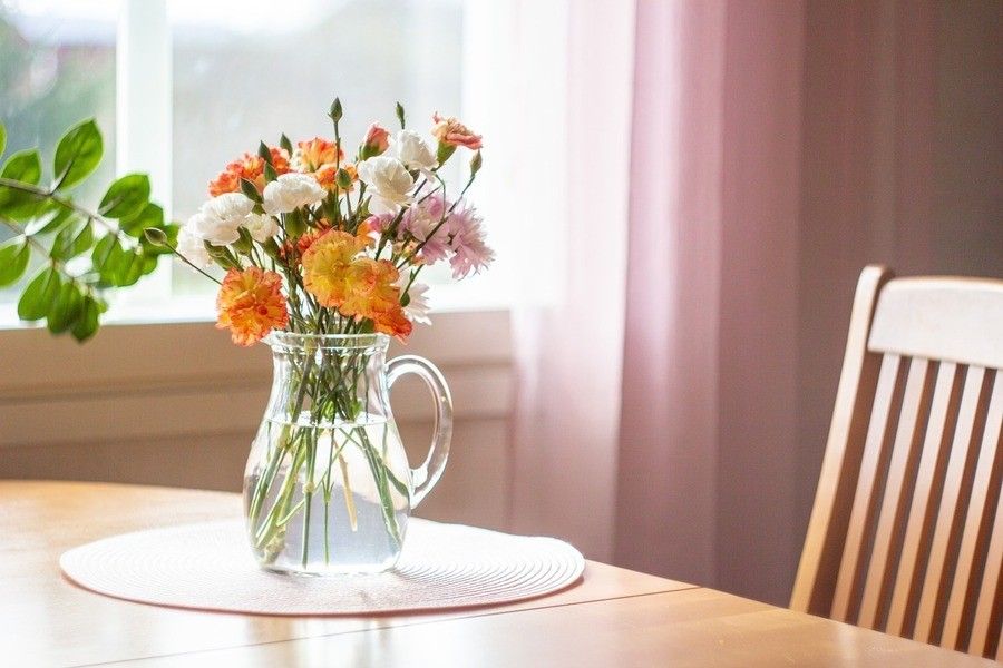 ¿Qué es un falso autónomo? Una jarra de cristal transparente llena de agua sostiene un ramo de flores rosas, blancas y naranjas sobre una mesa de madera cerca de una ventana con cortinas rosas transparentes, creando una escena serena que recuerda al acogedor espacio de trabajo de un falso autónomo. A la derecha se ve parcialmente una silla de madera.