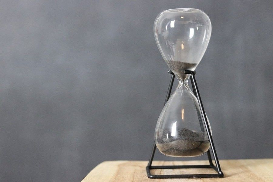 ¿Qué es un falso autónomo? Sobre una superficie de madera se coloca un reloj de arena negra, que recuerda las limitaciones de tiempo a las que se enfrenta un falso autónomo. Está sujeto por un marco de metal negro sobre un fondo gris liso.