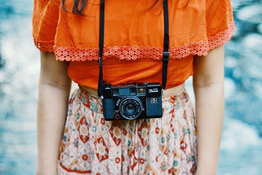 ¿Puede publicar mi ex fotos de mis hijos en internet sin mi consentimiento? Una persona con un top naranja y una falda estampada se sitúa delante de un fondo azul, con una cámara de cine vintage negra colgada del cuello por una correa, recordándonos que no debemos publicar imágenes de menores sin consentimiento.