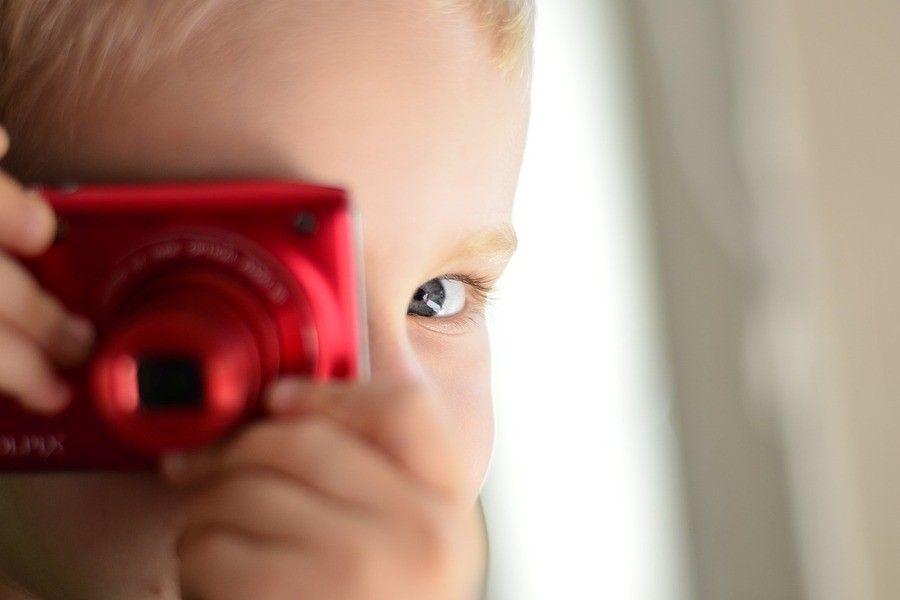 ¿Puede publicar mi ex fotos de mis hijos en internet sin mi consentimiento? Primer plano de un niño pequeño que sostiene una cámara digital roja en la cara, con un ojo visible mirando por encima de la cámara. El fondo está desenfocado, lo que subraya la importancia de no publicar imágenes de menores sin su consentimiento.