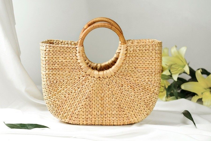 Un bolso de paja tejida con asas redondas está colocado sobre una superficie blanca. De fondo, lirios amarillos y hojas verdes que evocan la suave elegancia de un ama de casa. La iluminación es clara y natural. Un bolso de paja tejida con asas redondas está colocado sobre una superficie blanca. De fondo, lirios amarillos y hojas verdes que evocan la suave elegancia de un ama de casa. La iluminación es clara y natural.