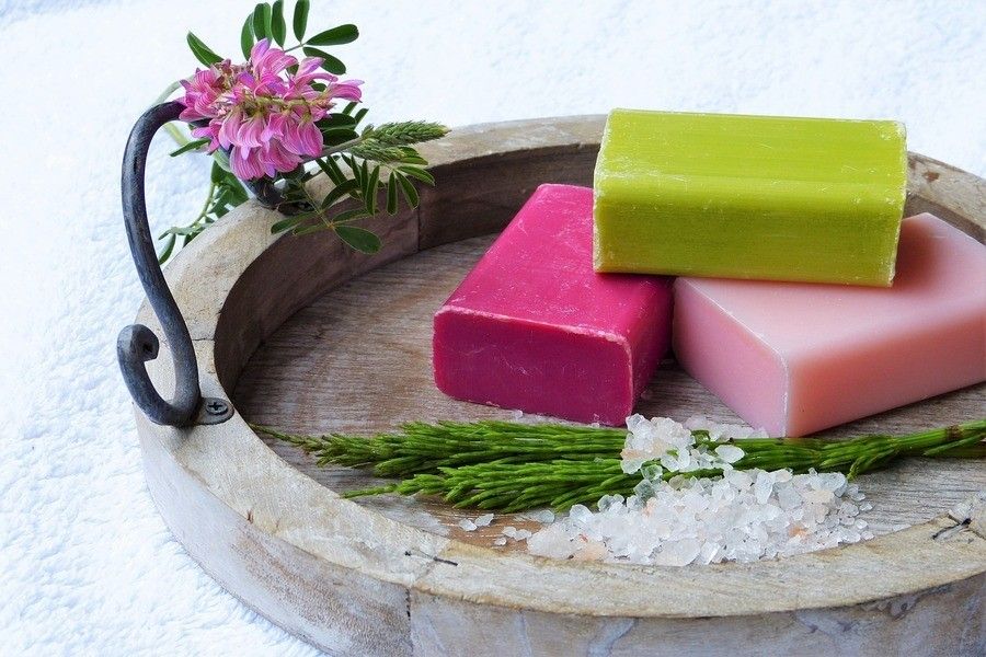 ¿Tengo derecho a pensión en caso de divorcio siendo ama de casa? Una bandeja redonda de madera contiene tres pastillas de jabón de colores -verde, rosa y rosa claro- junto a sal gruesa, ramitas de plantas verdes y una flor morada, evocando el tranquilo retiro que se merece un ama de casa tras una pensión de divorcio.