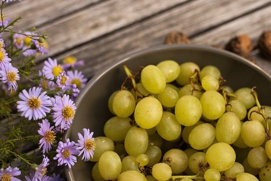 ¿Tengo derecho a pensión en caso de divorcio siendo ama de casa? Un cuenco lleno de uvas verdes descansa sobre una superficie de madera junto a un racimo de flores moradas y amarillas, evocando las sencillas comodidades de las que podría disfrutar un ama de casa. Al fondo se ven algunas nueces.