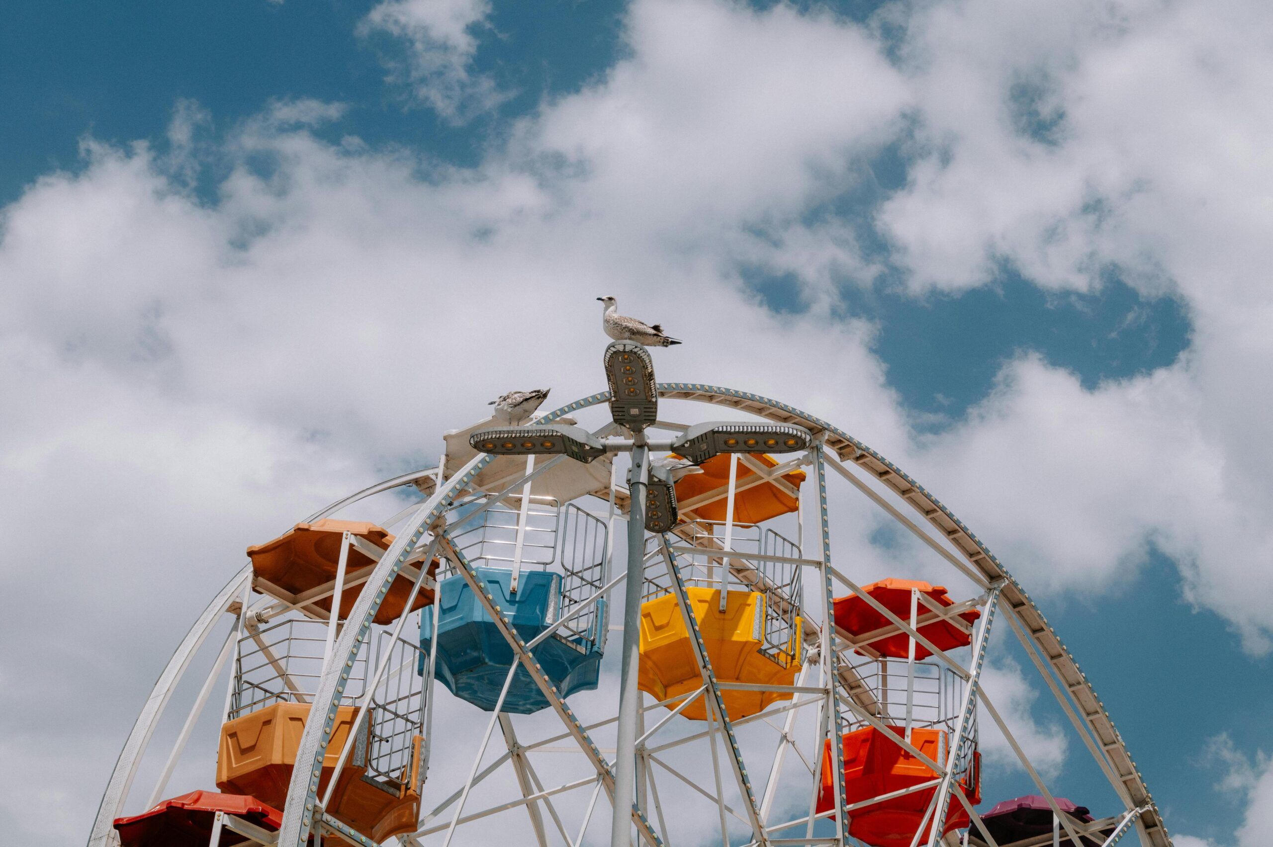 ¿Cómo es el reparto de vacaciones de verano en divorcios o separación? Una noria con góndolas rojas, amarillas y naranjas se alza sobre un cielo parcialmente nublado: dos gaviotas posadas en lo alto como si contemplaran el reparto de vacaciones de verano.