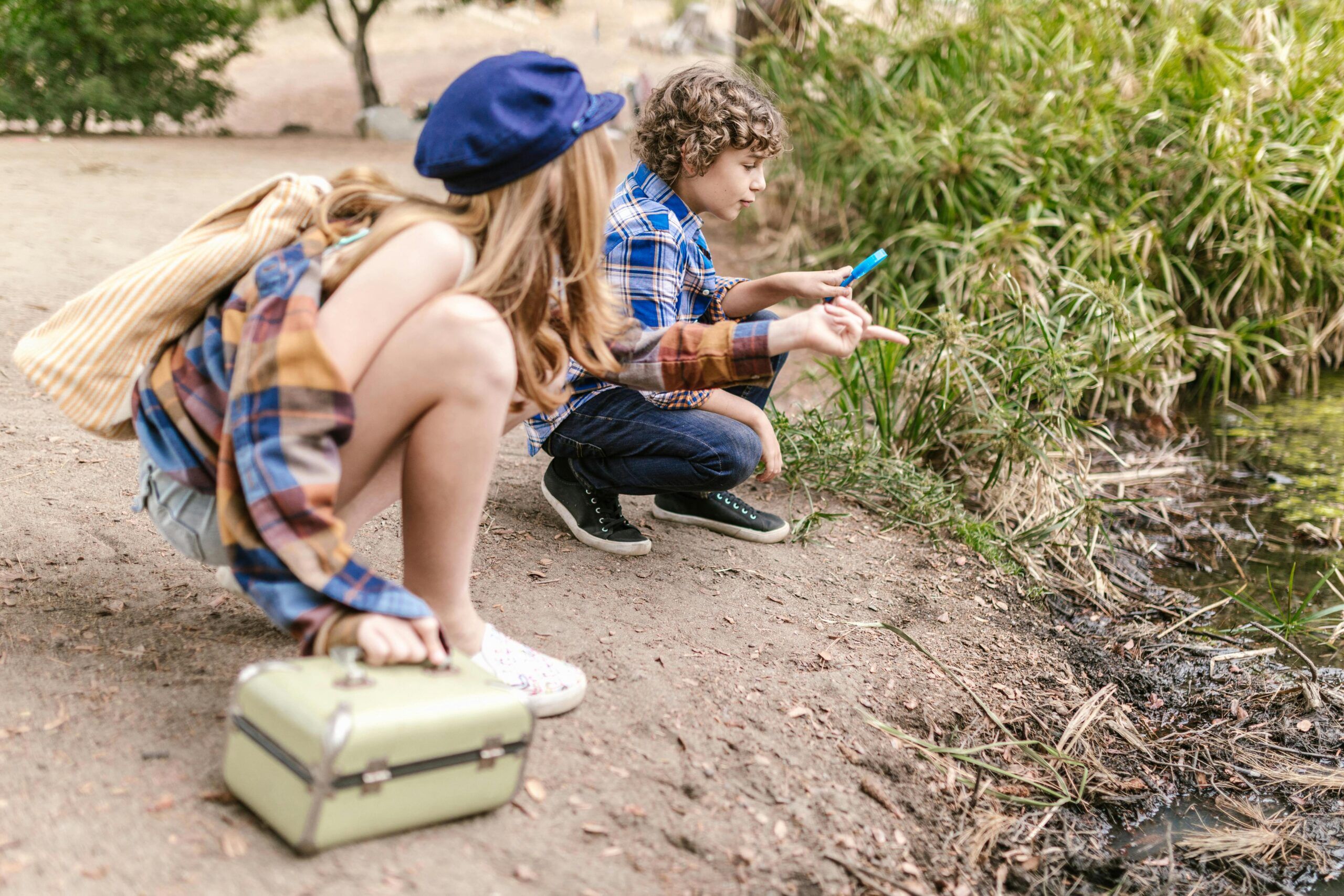 ¿Cómo es el reparto de vacaciones de verano en divorcios o separación? Dos niños se agazapan a la orilla del estanque, disfrutando de las vacaciones de verano. Uno sostiene una fiambrera verde claro; el otro utiliza una herramienta azul. Ambos llevan camisas de cuadros y ropa informal, rodeados de vegetación: un momento de paz durante el reparto de vacaciones de verano tras un divorcio.