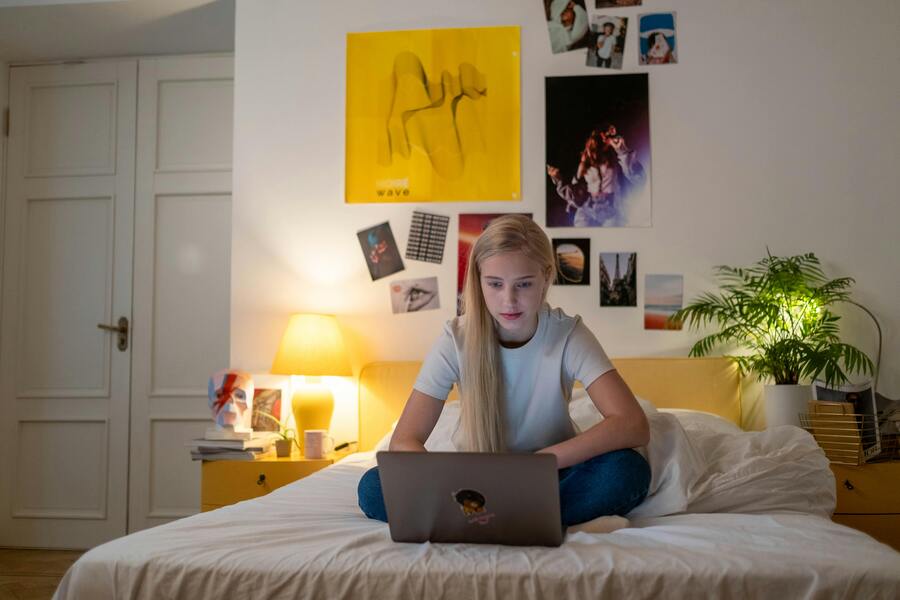 ¿Cómo puedo adoptar al hijo de mi pareja? Un joven se sienta con las piernas cruzadas en una cama y utiliza un ordenador portátil en un dormitorio con carteles en la pared, quizás investigando temas como adoptar al hijo de mi pareja, con una lámpara en la mesilla de noche y una maceta cerca.