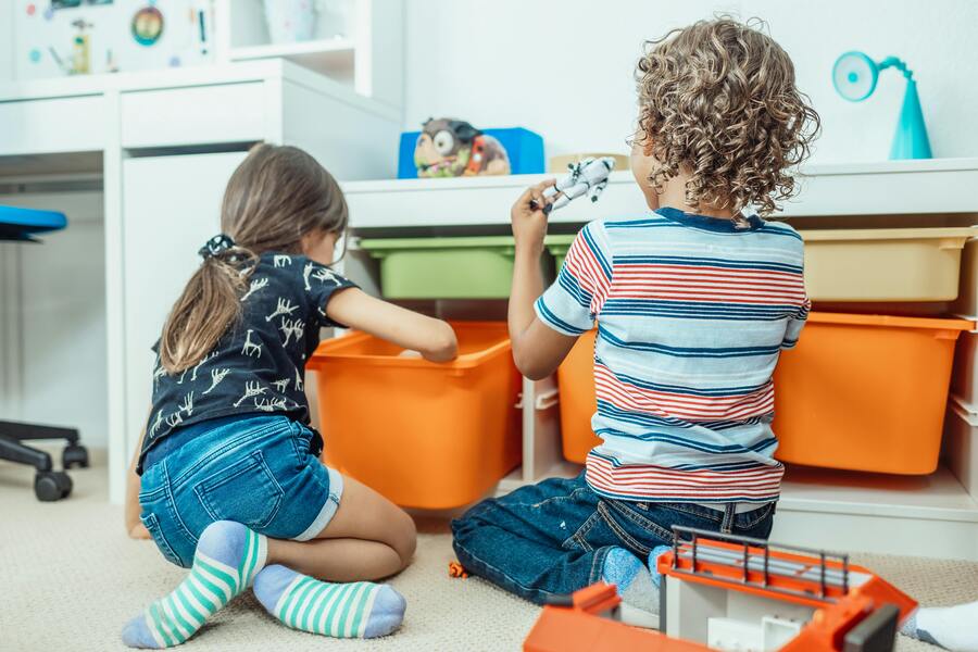 ¿Cómo puedo adoptar al hijo de mi pareja? Dos niños sentados en el suelo, de espaldas, organizan cubos naranjas bajo una mesa blanca en una habitación luminosa. Momentos como estos pueden acercar a las familias, sobre todo a las que se plantean adoptar al hijo de mi pareja.