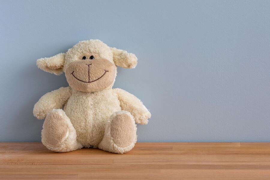 Una oveja de peluche con una gran sonrisa cosida está sentada sobre una superficie de madera contra una pared azul claro. Una oveja de peluche con una gran sonrisa cosida está sentada sobre una superficie de madera contra una pared azul claro.