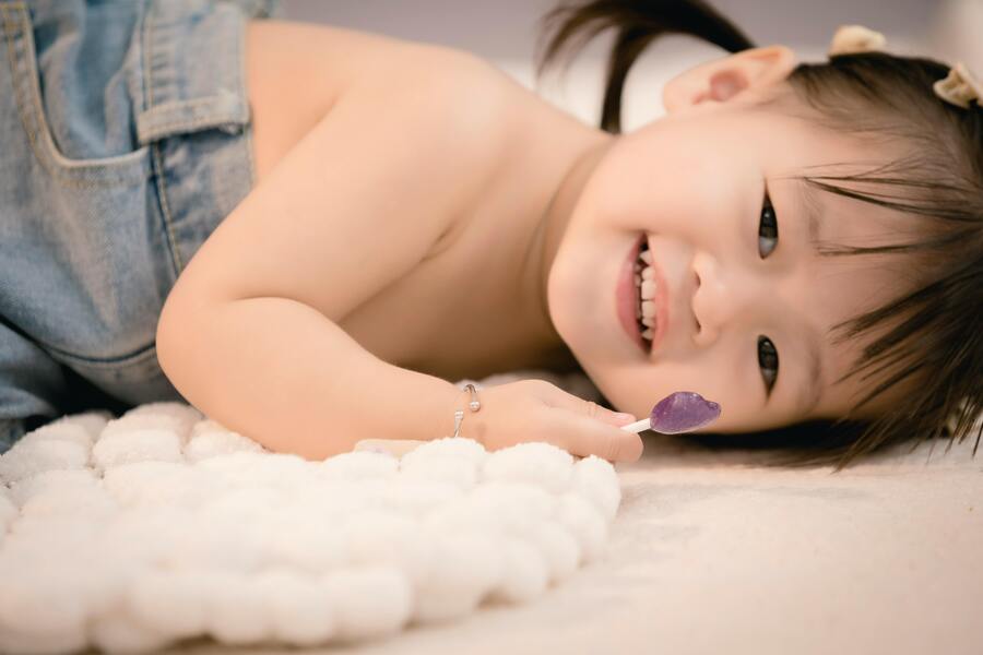 Qué es la custodia exclusiva o monoparental Una niña pequeña con el pelo oscuro recogido en coletas está tumbada sobre una superficie de textura suave y blanca, sonriendo y sosteniendo una piruleta morada cerca de la cara. Lleva un mono vaquero con un tirante fuera del hombro.