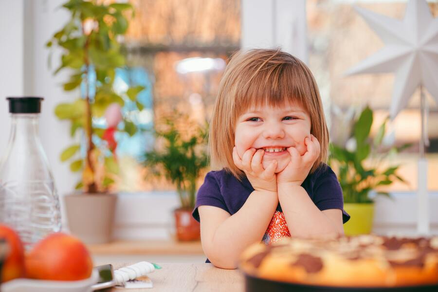Qué es la custodia exclusiva o monoparental Un niño pequeño de pelo corto castaño claro sonríe con las manos apoyadas en las mejillas, sentado en una mesa con fruta y un pastel en una habitación luminosa con plantas y una gran estrella de papel como decoración.