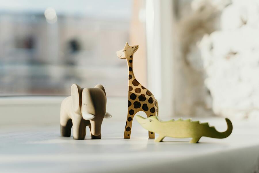 Tres figuras de madera de animales -un elefante, una jirafa con manchas y un cocodrilo verde- se colocan sobre una superficie blanca cerca de una ventana con luz natural suave. Tres figuras de madera de animales -un elefante, una jirafa con manchas y un cocodrilo verde- se colocan sobre una superficie blanca cerca de una ventana con luz natural suave.