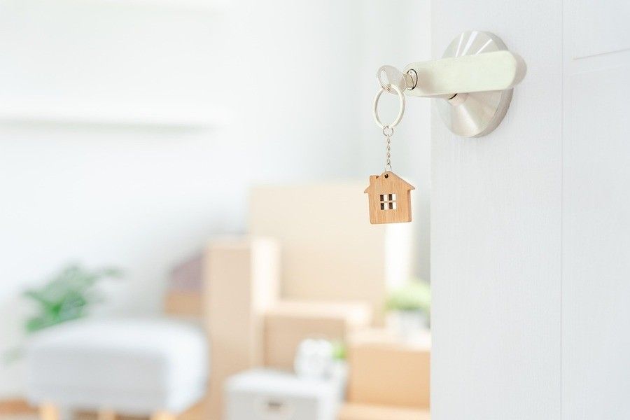 Primer plano de una puerta con picaporte plateado, que muestra una llave con un llavero en forma de casa insertado en la cerradura. En el fondo borroso, hay cajas de cartón y muebles, lo que sugiere una escena de mudanza. Primer plano de una puerta con picaporte plateado, que muestra una llave con un llavero en forma de casa insertado en la cerradura. En el fondo borroso, hay cajas de cartón y muebles, lo que sugiere una escena de mudanza.