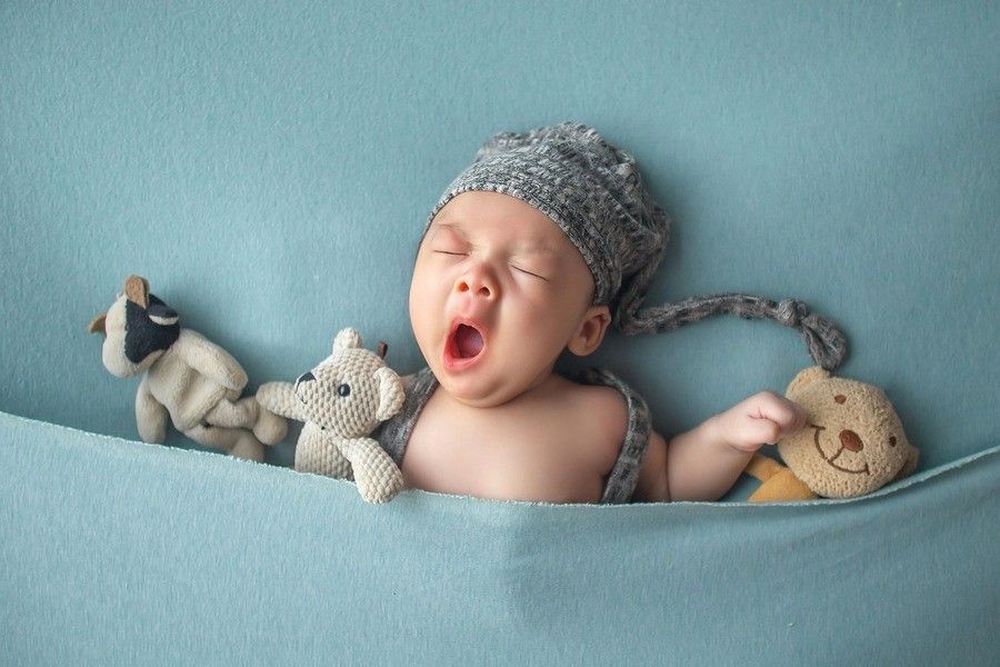 Un bebé con un gorro de punto gris está tumbado sobre una manta azul, bosteza y sostiene tres peluches. El bebé parece estar parcialmente metido bajo la manta.