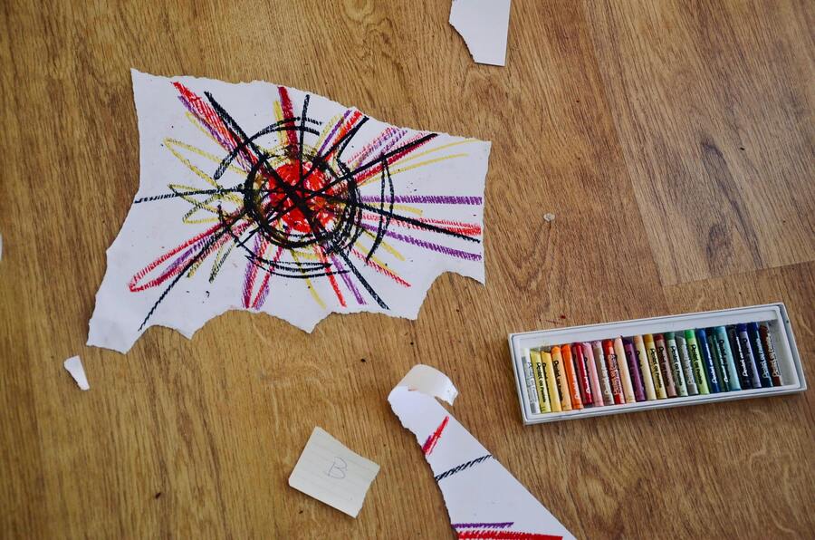 En el suelo de madera hay un trozo de papel roto con un colorido dibujo abstracto hecho con ceras de colores. Cerca, hay una caja de ceras de colores variadas y más trozos de papel rotos esparcidos por los alrededores.