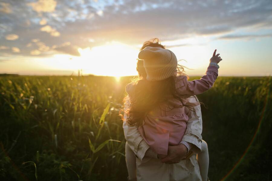 Un adulto lleva en brazos a un niño en un campo durante la puesta de sol. El niño lleva un sombrero claro y una chaqueta rosa y señala hacia el cielo. El cielo está parcialmente nublado y brilla la luz del sol. Ambos son vistos desde atrás. Un adulto lleva en brazos a un niño en un campo durante la puesta de sol. El niño lleva un sombrero claro y una chaqueta rosa y señala hacia el cielo. El cielo está parcialmente nublado y brilla la luz del sol. Ambos son vistos desde atrás.