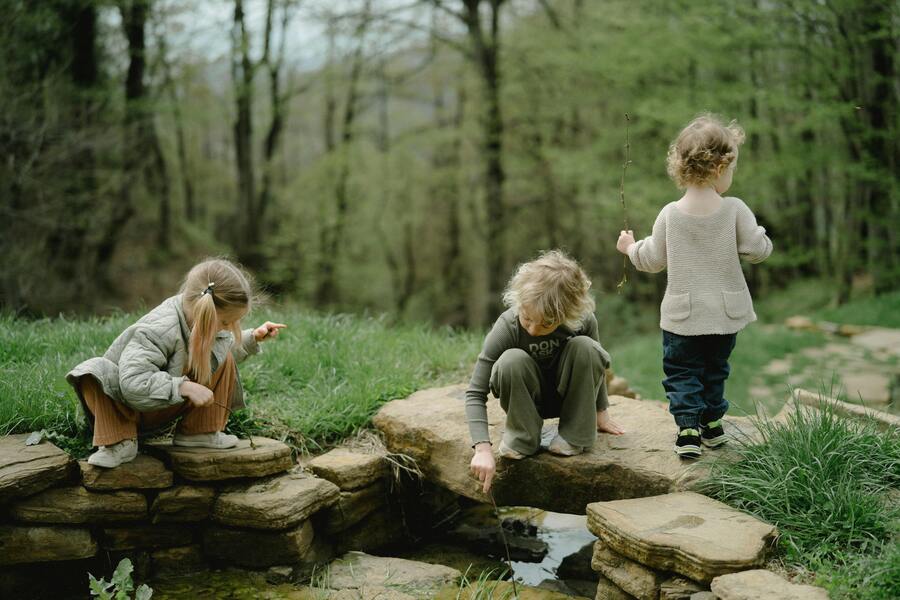 Tres niños pequeños juegan al aire libre junto a un pequeño arroyo bordeado de piedras, rodeado de hierba verde y árboles. Dos de ellos están agachados junto al agua, mientras que otro sujeta un palo. La escena parece tranquila y natural. Tres niños pequeños juegan al aire libre junto a un pequeño arroyo bordeado de piedras, rodeado de hierba verde y árboles. Dos de ellos están agachados junto al agua, mientras que otro sujeta un palo. La escena parece tranquila y natural.