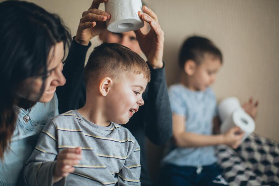Dos adultos y dos niños pequeños están en el interior de una casa. Un adulto sostiene un rollo de papel higiénico por encima de la cabeza de un niño, mientras que el otro niño, al fondo, sostiene un rollo de papel higiénico en las manos. La escena parece alegre.