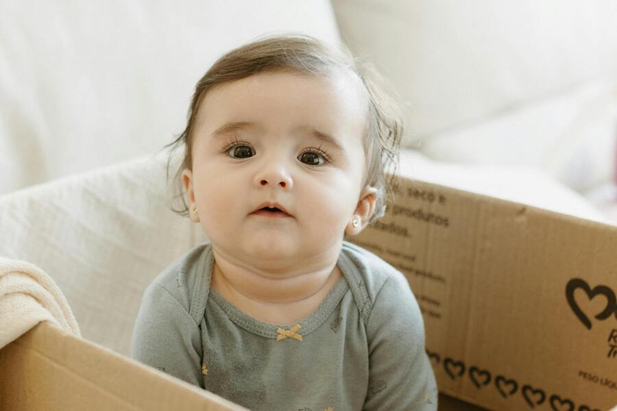 Un bebé de pelo castaño y con un body azul claro está sentado dentro de una caja de cartón, mirando ligeramente hacia arriba. El fondo incluye un sofá beige y parte de una manta.