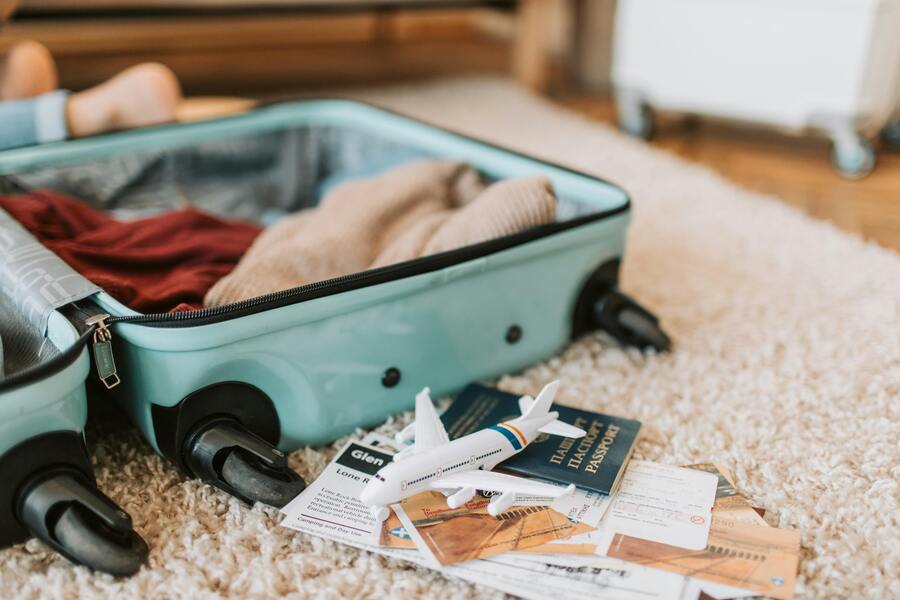 Sobre una alfombra hay una maleta abierta con ropa en su interior. Delante de la maleta hay documentos de viaje, un avión en miniatura y un pasaporte. La escena sugiere los preparativos de un viaje. Sobre una alfombra hay una maleta abierta con ropa en su interior. Delante de la maleta hay documentos de viaje, un avión en miniatura y un pasaporte. La escena sugiere los preparativos de un viaje.
